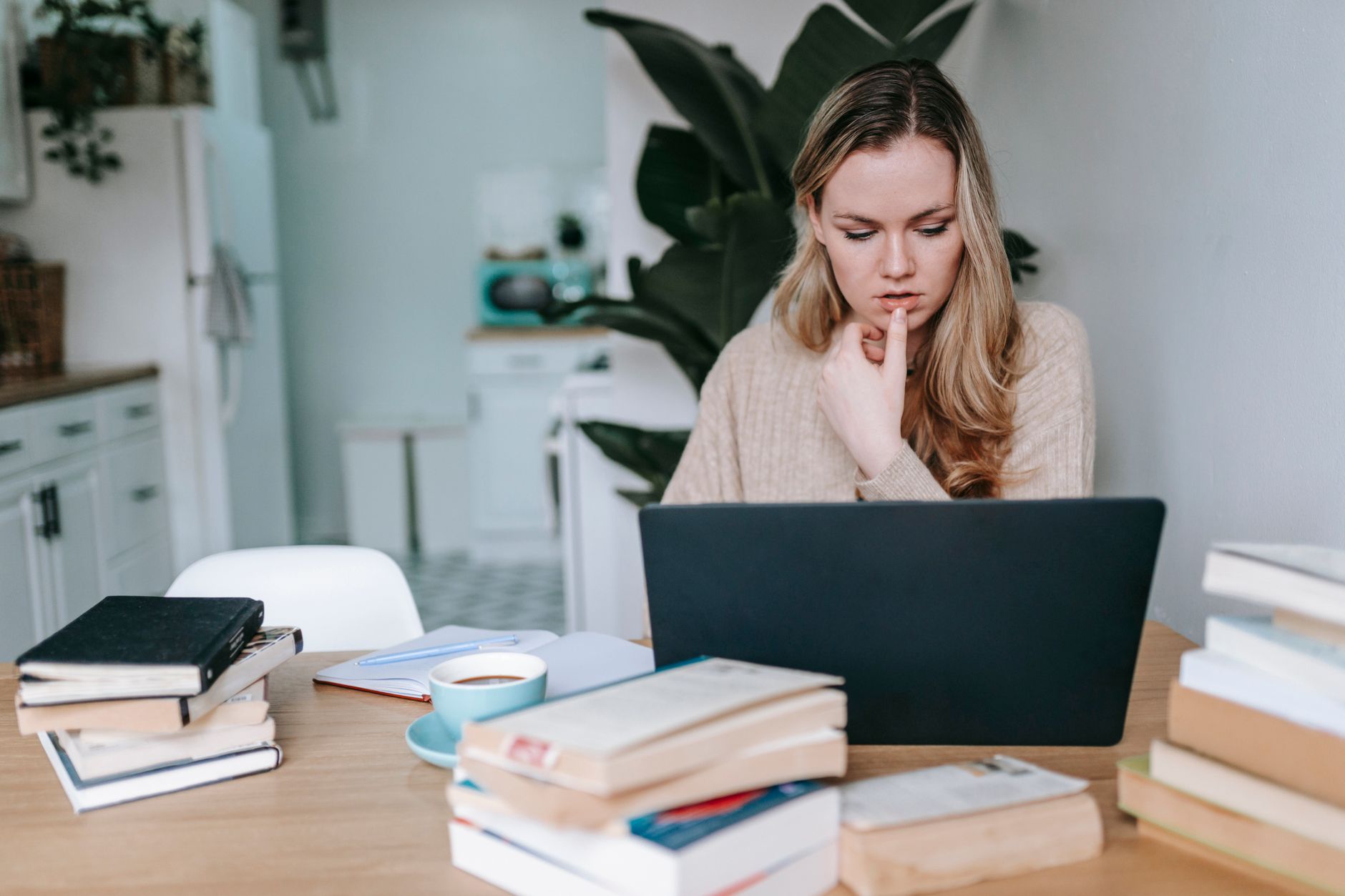 A woman studies at a laptop at her kitchen table, surrounded by stacks of books and a cup of coffee