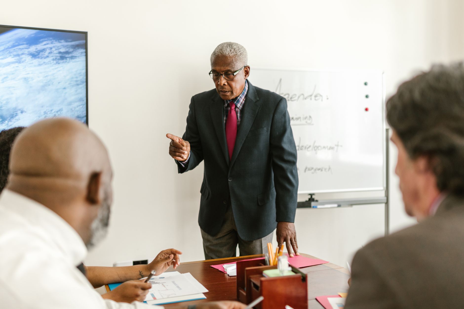 An older agency leader in a suit stands at the head of a conference table, leading a meeting with colleagues beside a whiteboard agenda