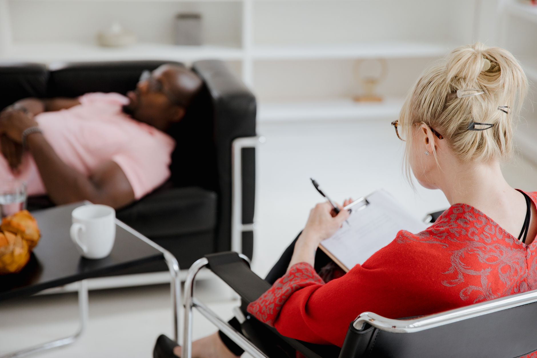 A therapist in a red sweater takes notes on a clipboard while a client rests on a leather sofa in a bright office