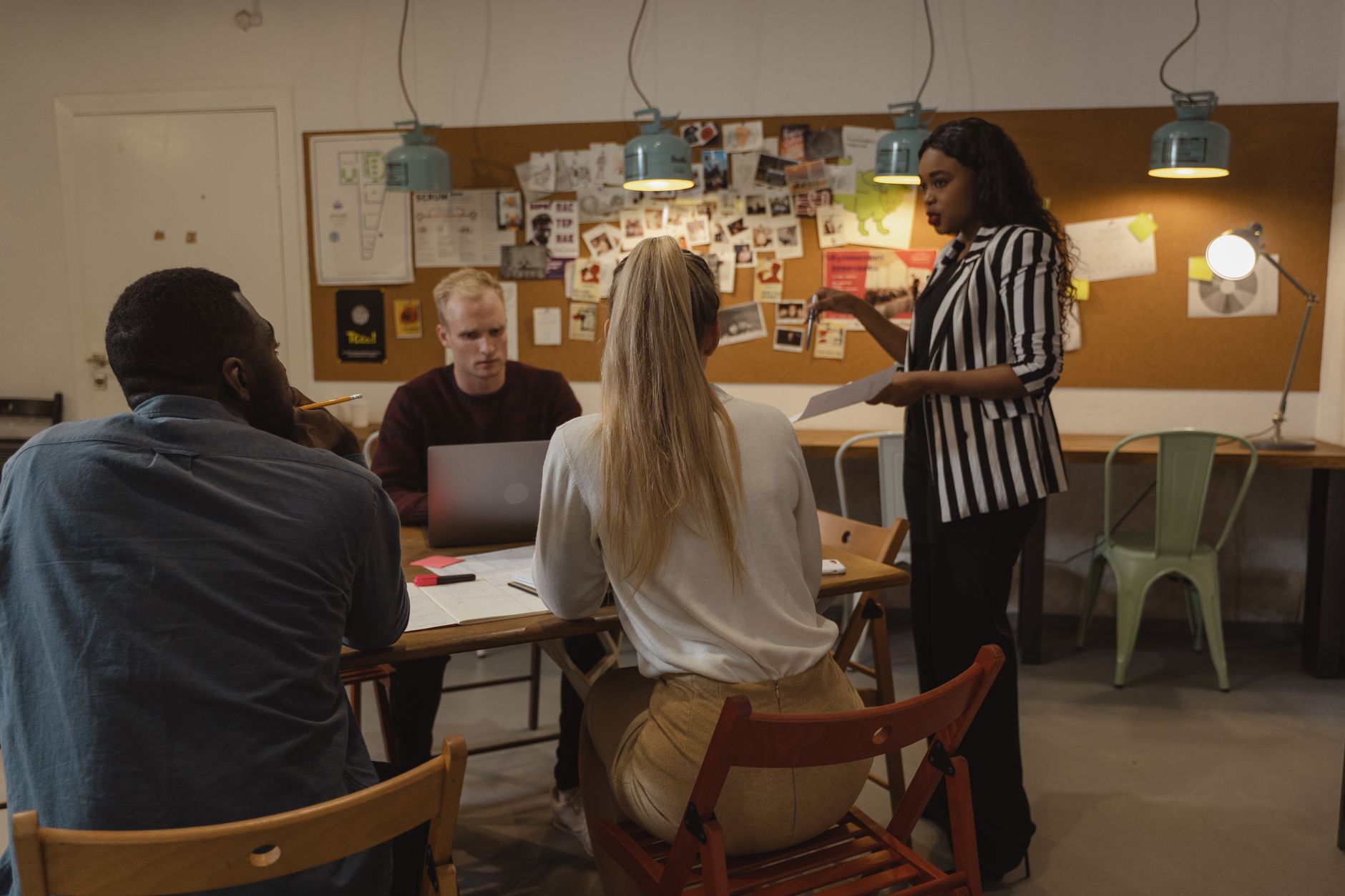 A woman in a striped blazer leads a team discussion at a wooden table while three colleagues listen and take notes