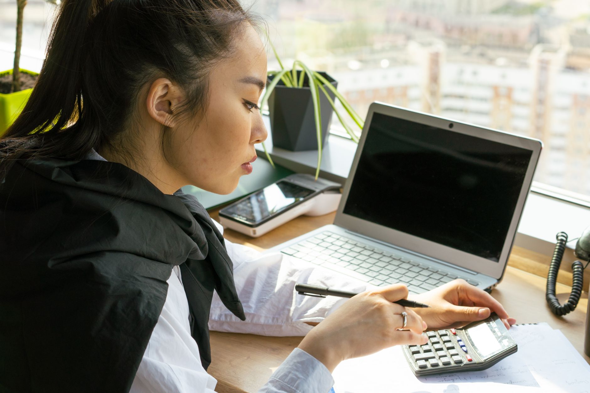 A woman works through figures on a calculator beside her laptop at a desk by a window