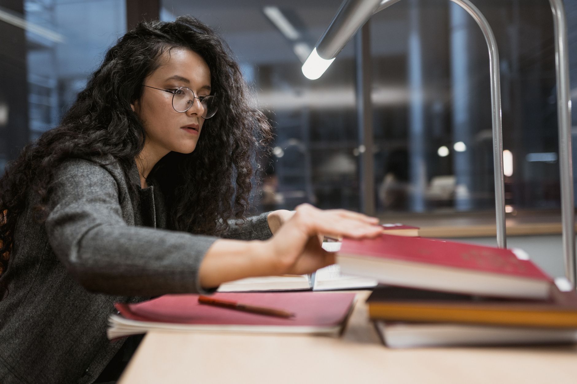 A doctoral student in glasses reaches for a book at a lamp-lit library desk stacked with research volumes