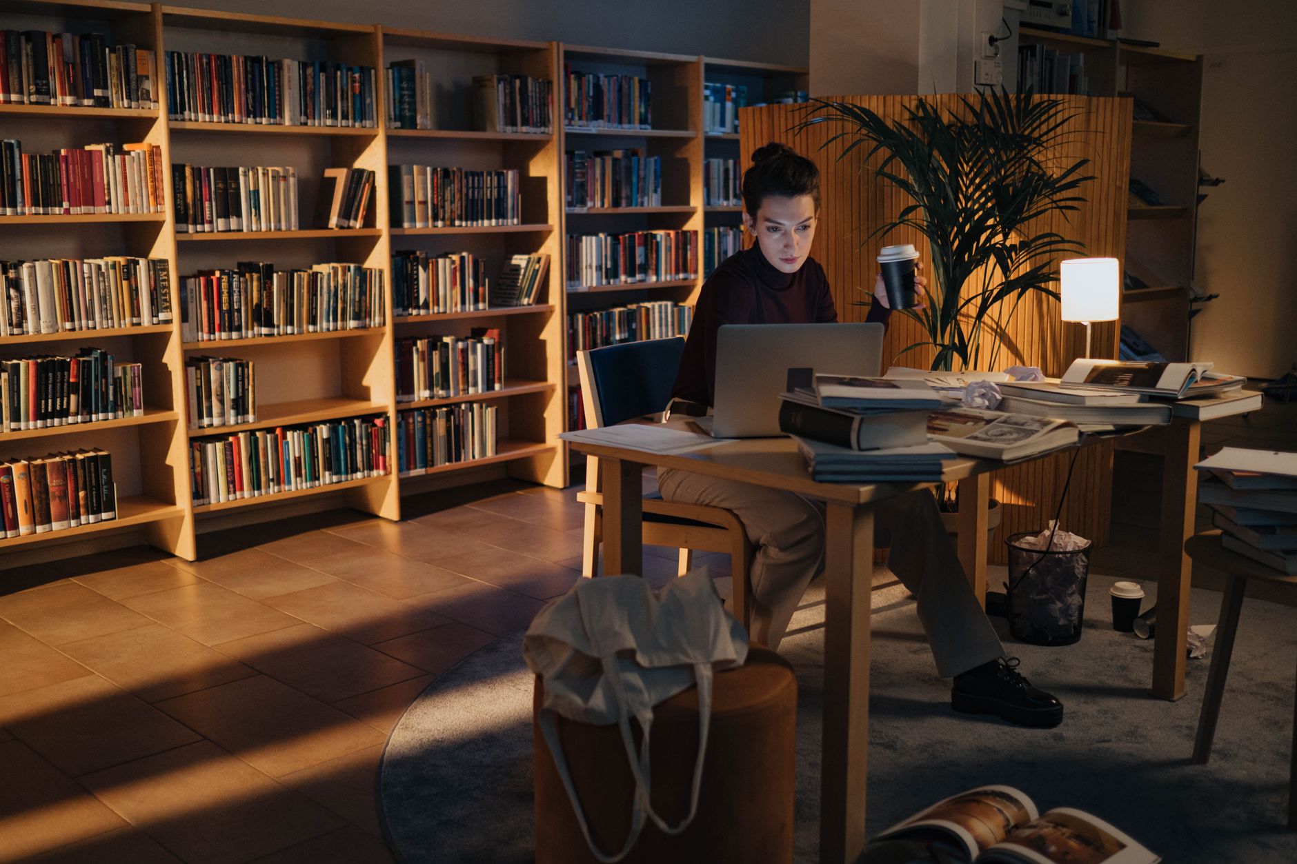 A woman studies late at a library table with a laptop, coffee and stacks of open books