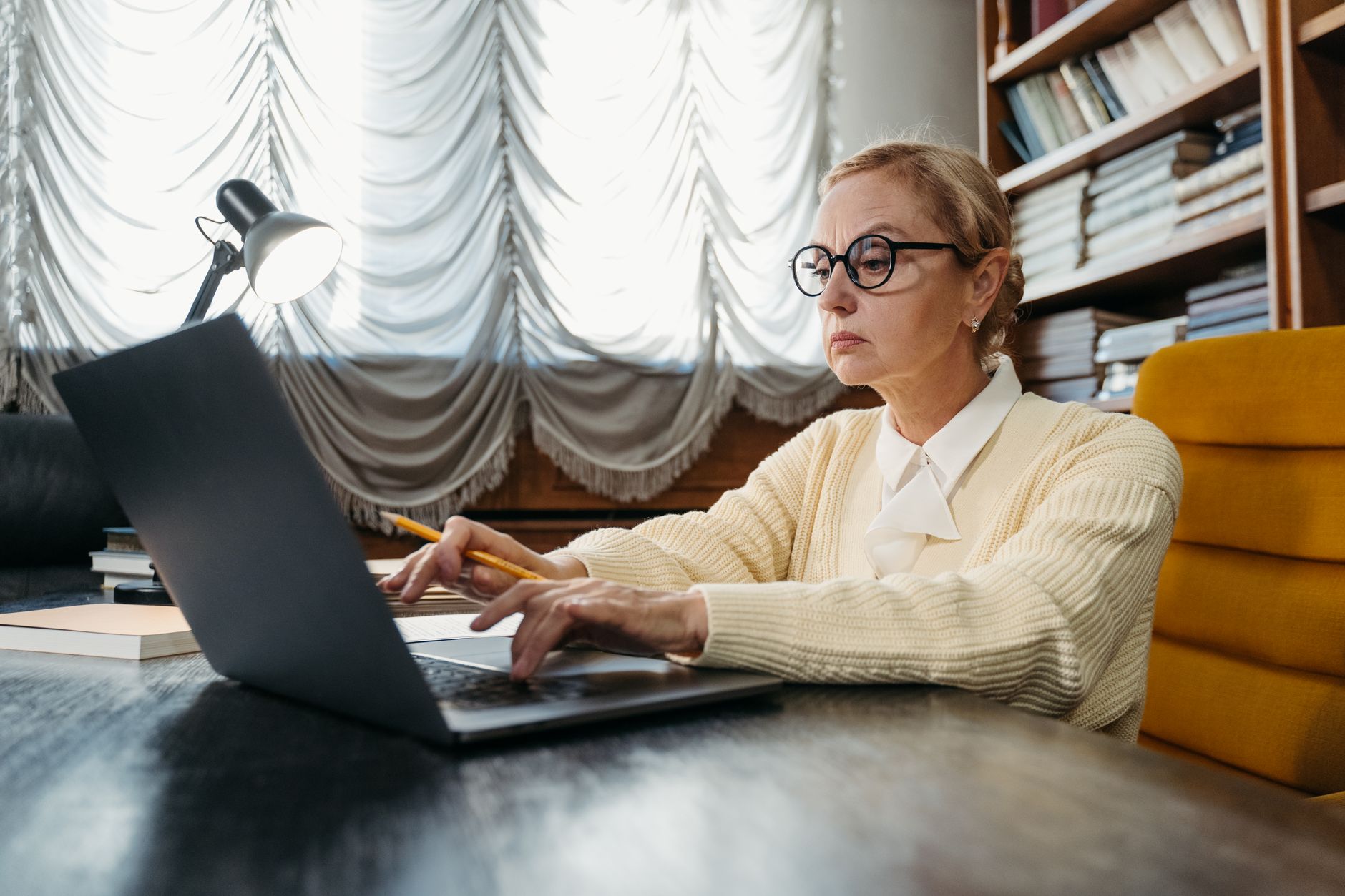 A social worker in glasses types on a laptop at a desk lined with bookshelves, a pencil in hand