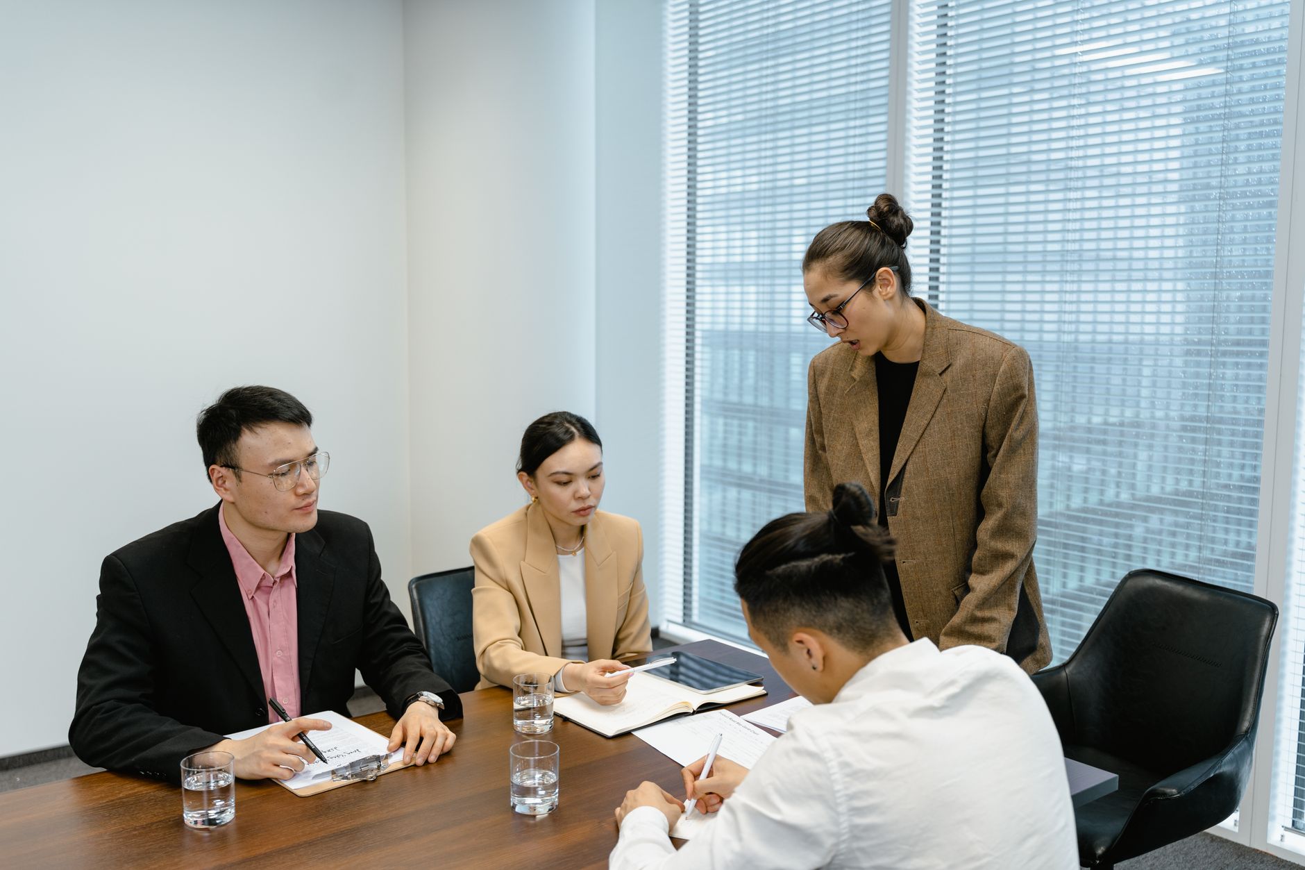 A woman in a tweed blazer stands and leads a planning meeting with three colleagues seated at a conference table