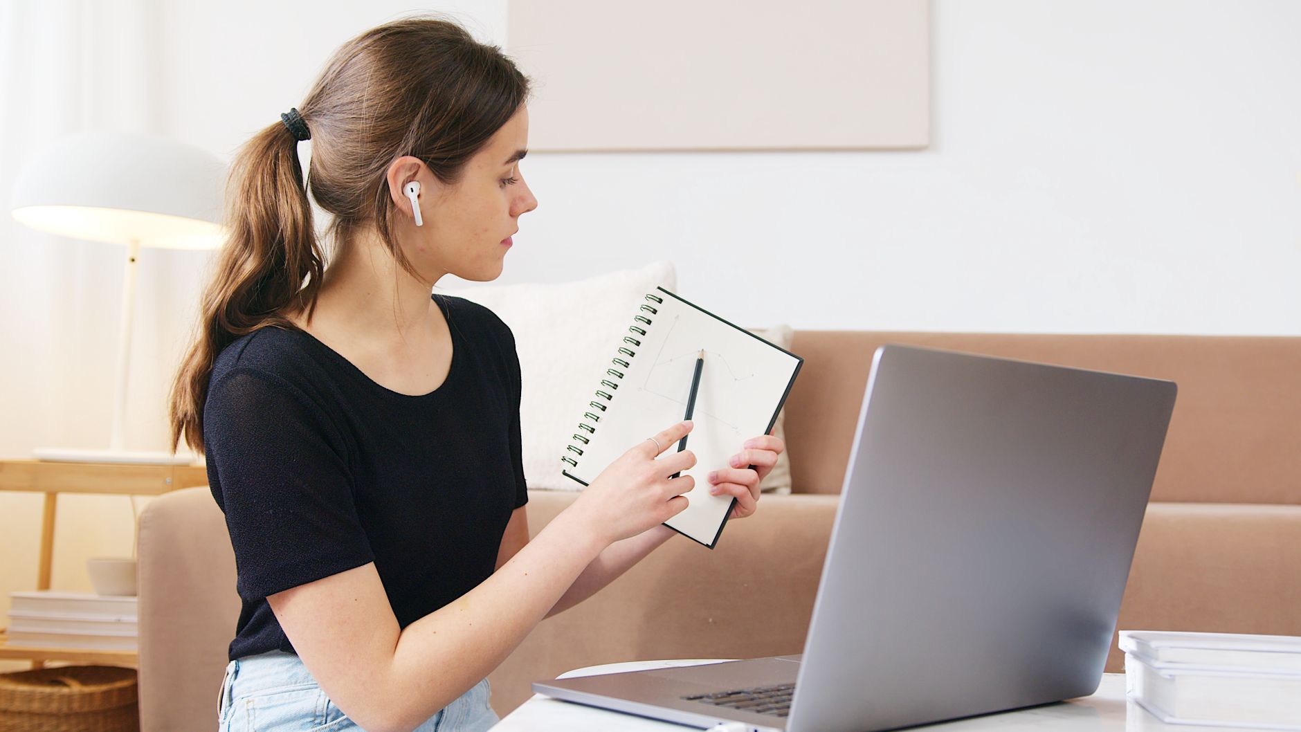 A doctoral student wearing earbuds holds up a notebook chart during a live video class on her laptop at home