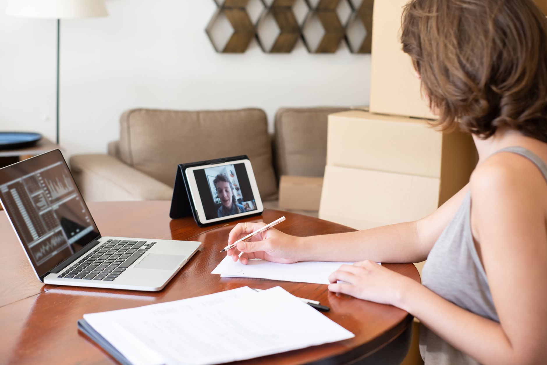 A social worker takes notes at her dining table while a classmate speaks on a tablet video call beside her laptop