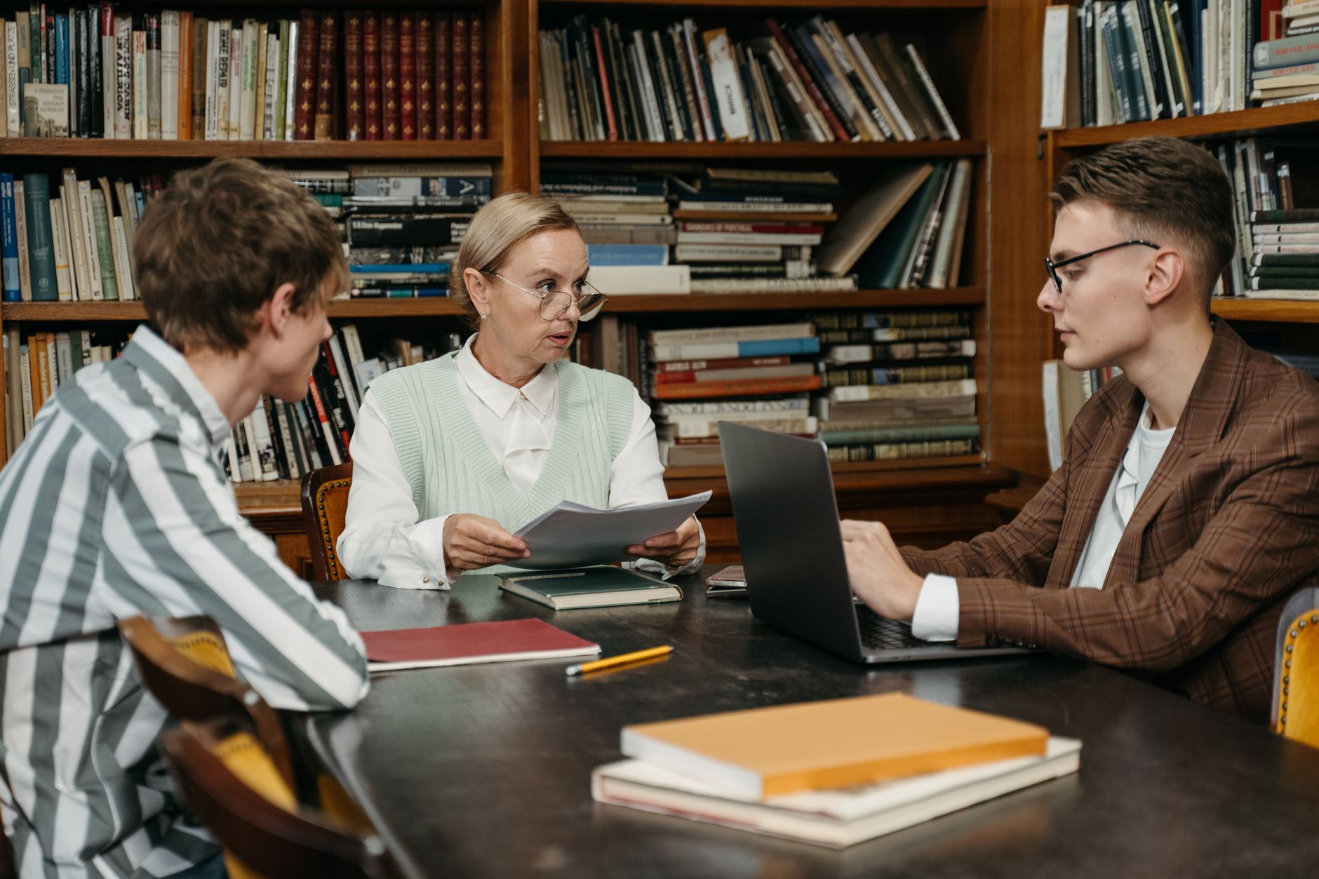 A professor reviews a paper with two doctoral students at a library table lined with bookshelves