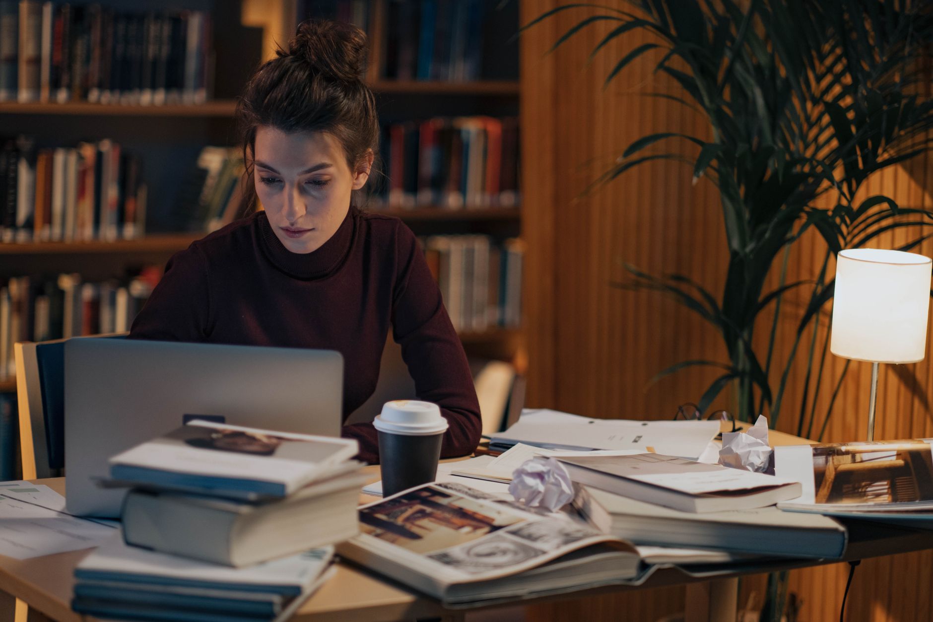 A woman works on a laptop at a library table stacked with open books and papers, with bookshelves behind her